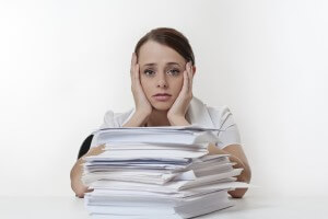 A stressed female with her head in her hands, sitting at her desk with a large pile of paper work stacked in front of her.
